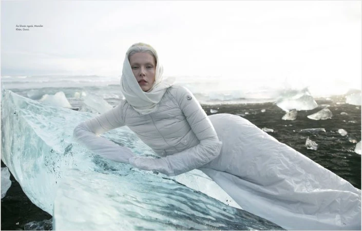 A woman in a white outfit poses on ice in an outdoor environment, captured in a fashion-style photograph with muted tones and natural light.