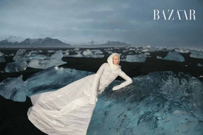 Fashion shot of a model in white dress and head covering reclining on an ice floe in a black sand environment with mountains in the background.