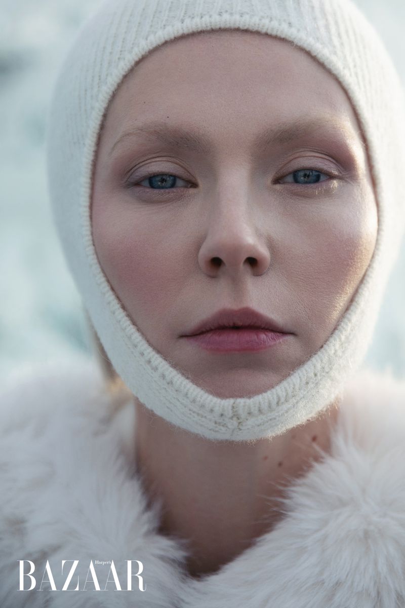 Close-up studio shot of a fair-skinned adult female model with blue eyes wearing a white knit balaclava and a white faux fur coat.