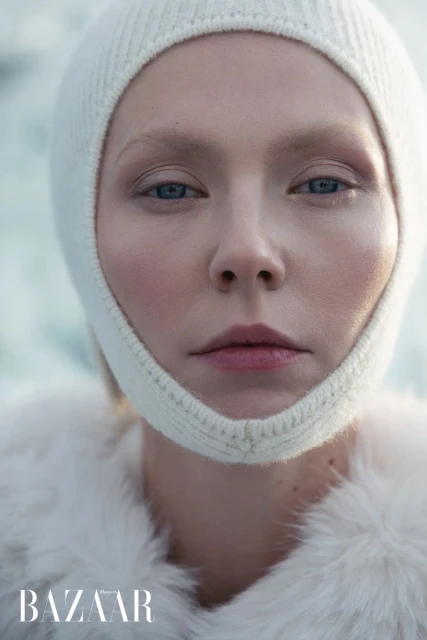 Close-up studio shot of a fair-skinned adult female model with blue eyes wearing a white knit balaclava and a white faux fur coat.