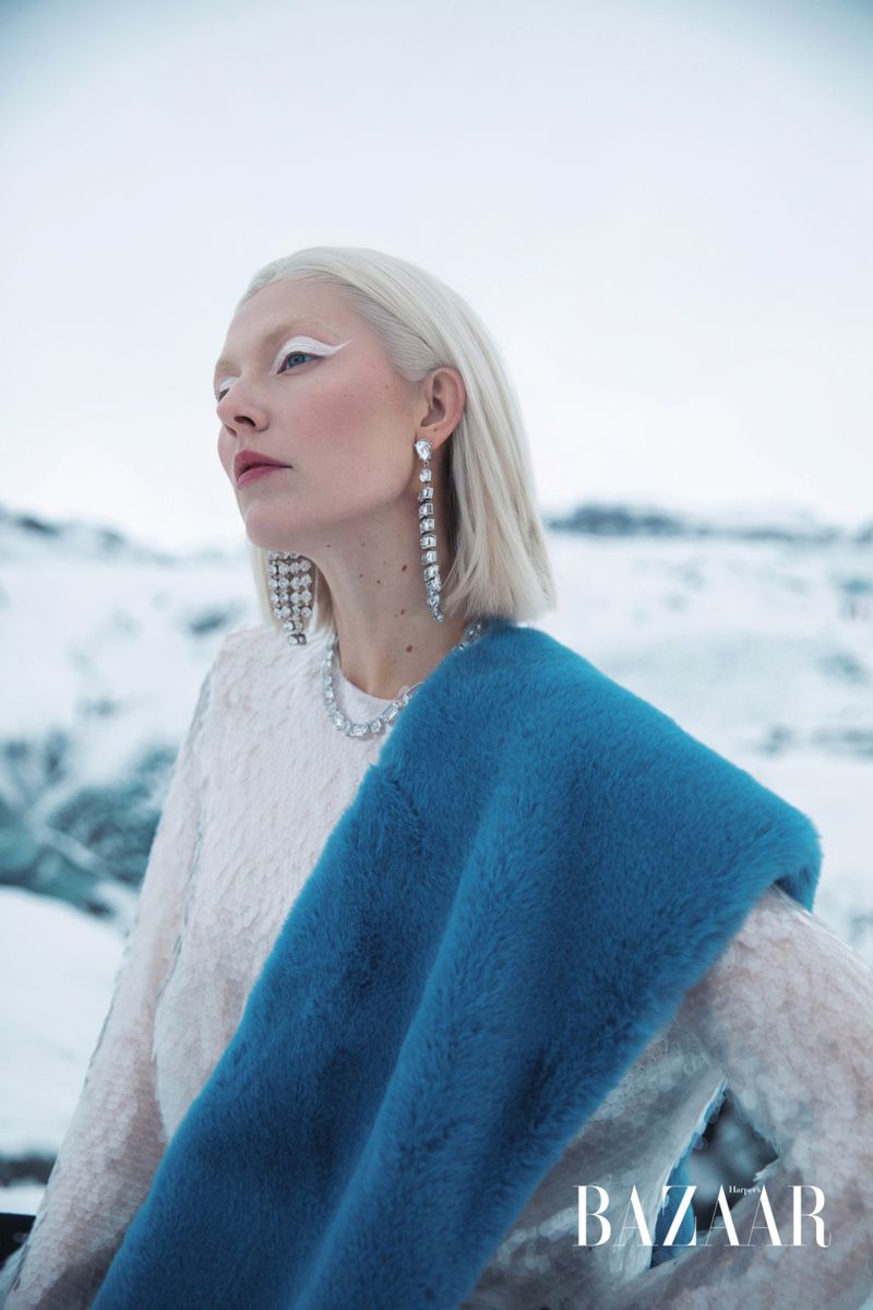 A woman with platinum hair, a bright blue stole, and distinctive white eyeliner poses in a snowy, mountainous landscape. Fashion shot.
