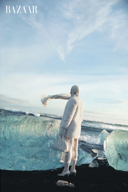 Fashion editorial shot of a woman in a white outfit, wearing a hood, holding a purse, standing on black sand, with icebergs in Iceland.