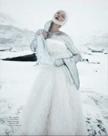 Model in white feathery gown and silver sequined top wearing a matching white head covering in a snowy landscape.