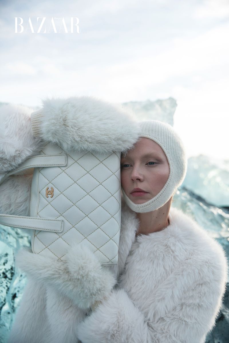 Editorial fashion shot features a woman in a fur coat and knit balaclava holding a quilted handbag in front of a glacier.