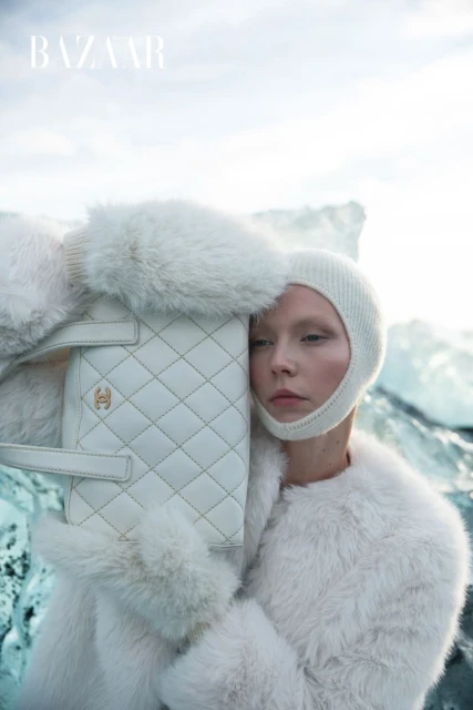 Editorial fashion shot features a woman in a fur coat and knit balaclava holding a quilted handbag in front of a glacier.