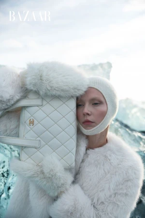 Editorial fashion shot features a woman in a fur coat and knit balaclava holding a quilted handbag in front of a glacier.