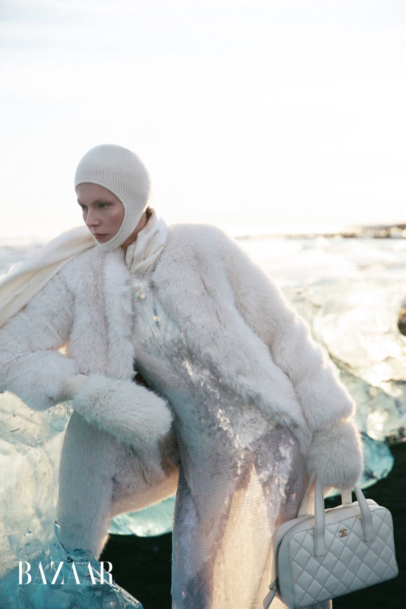 Fashion editorial shot of a pale woman in all-white winter clothing with a handbag, standing on ice near a bright sky.
