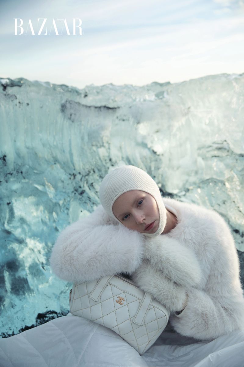 Fashion shot of a woman in white faux fur coat and knit hat with a quilted handbag in front of an icy glacier and light blue sky.