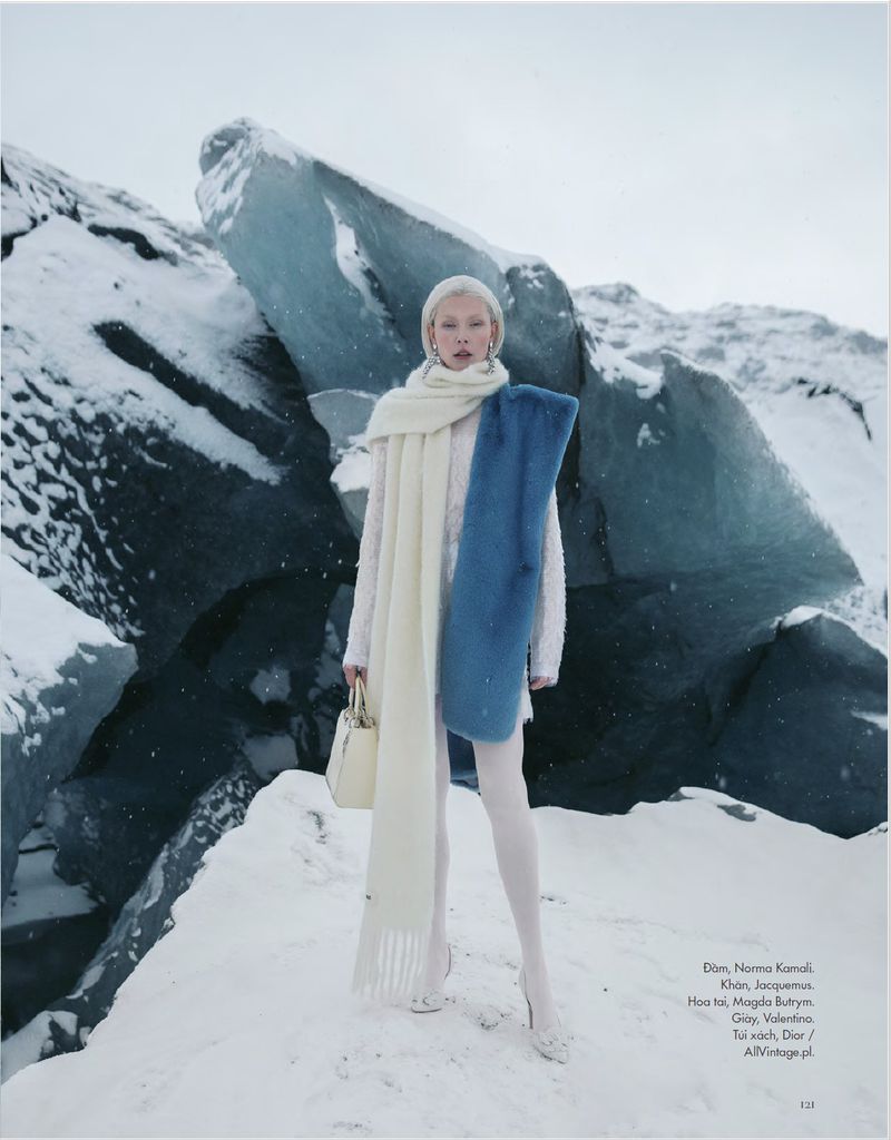Fashion model in an all-white outfit poses with a handbag and long scarf in a snowy landscape with ice formations.