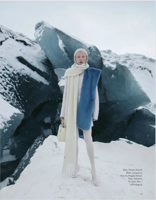 Fashion model in an all-white outfit poses with a handbag and long scarf in a snowy landscape with ice formations.