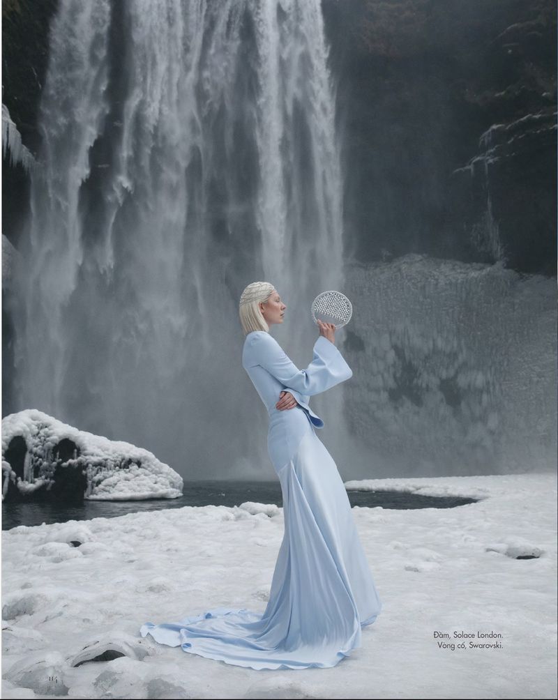 Fashion shot of a fair-skinned woman in a light-blue dress, holding a silver sphere, with a large waterfall and snowy landscape behind her.