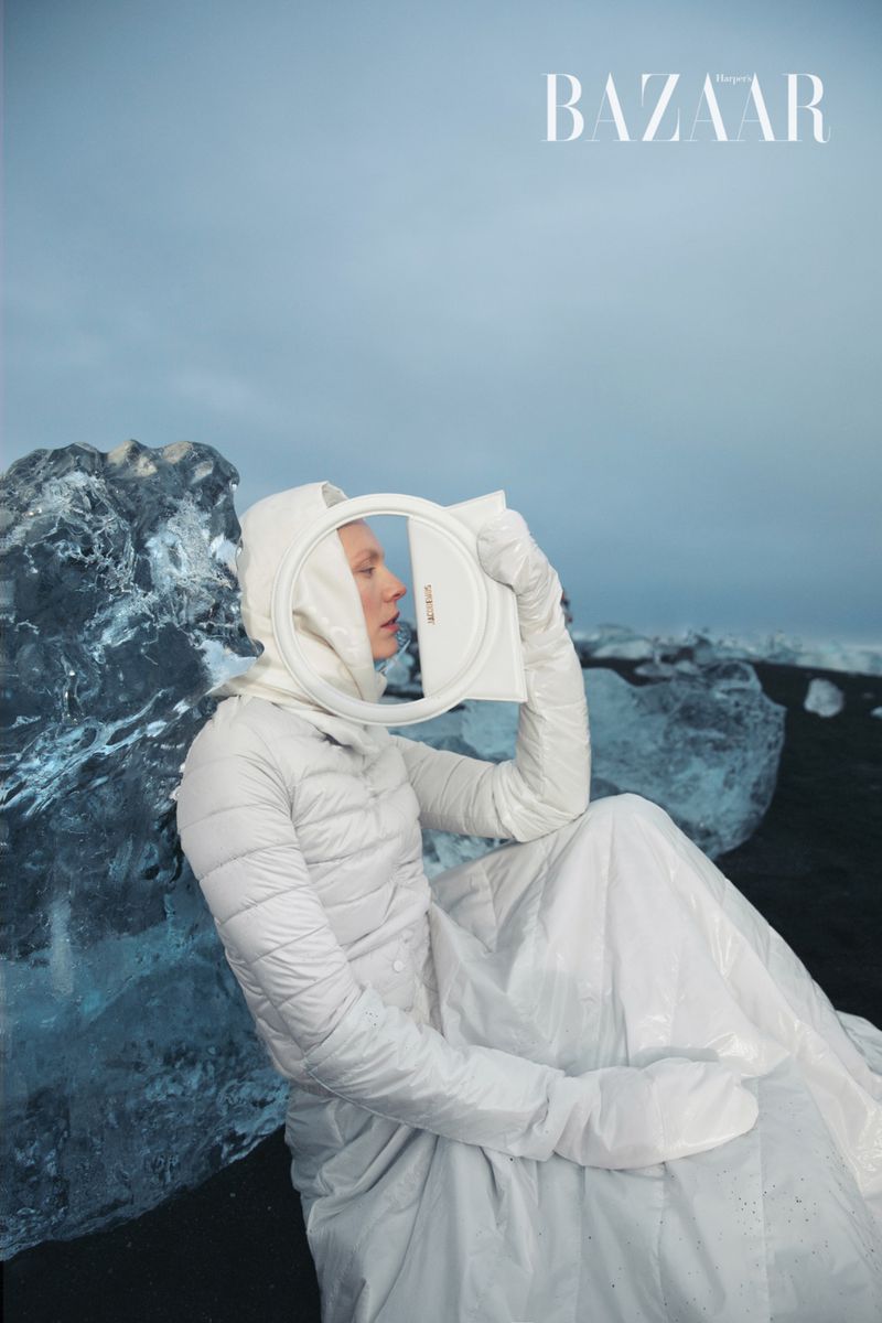 Fashion shot of a fair-skinned model in white hooded jacket, skirt, and gloves, holding a white framed object on a black sand beach.