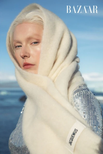Close-up of a fair-skinned woman wearing a silver sequin top and a cream-colored scarf. She poses outdoors in front of a body of water.