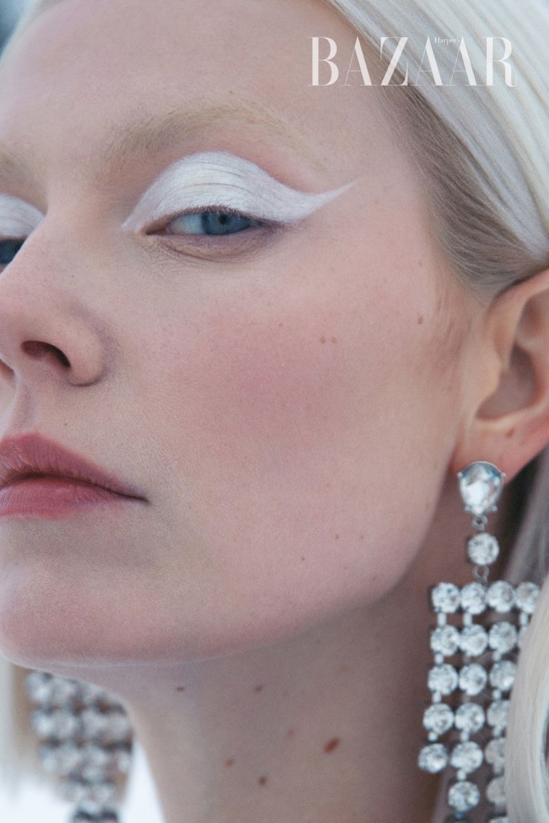 Close-up of a woman with fair skin and platinum blonde hair showcasing striking white eyeshadow and chandelier earrings, with 'BAZAAR' text overlay.