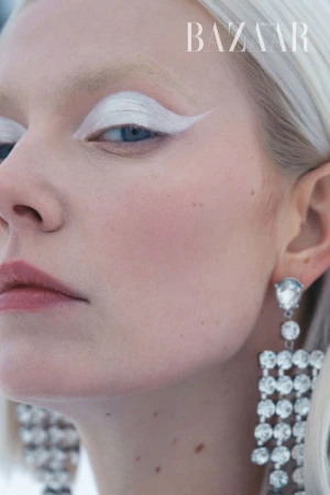 Close-up of a woman with fair skin and platinum blonde hair showcasing striking white eyeshadow and chandelier earrings, with 'BAZAAR' text overlay.