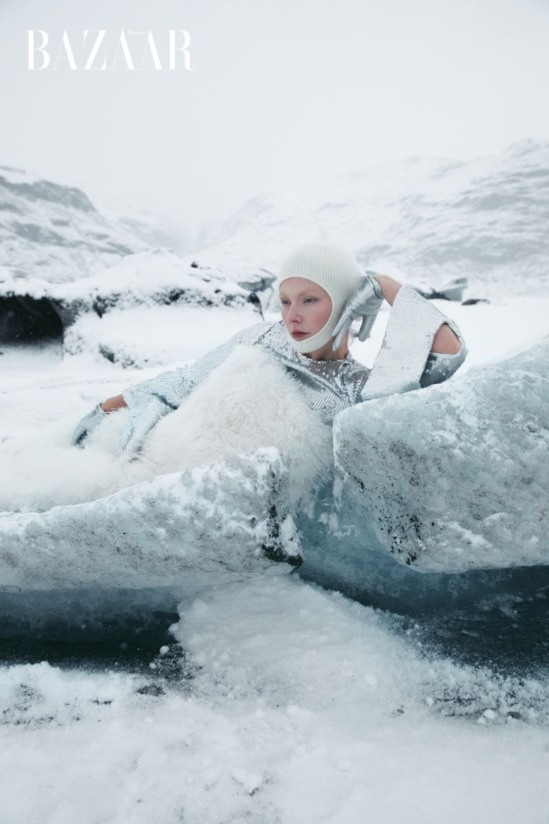 Fashion editorial featuring a woman in a shiny silver outfit and head covering, posing in an icy, snowy landscape with mountains in the background.