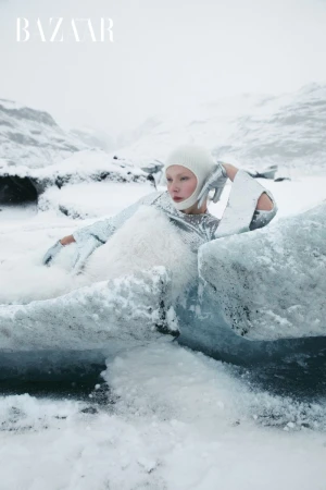 Fashion editorial featuring a woman in a shiny silver outfit and head covering, posing in an icy, snowy landscape with mountains in the background.