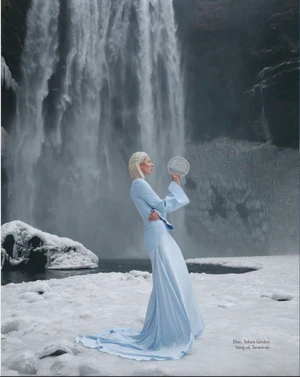 Model in a pale blue gown holding a circular prop before a frozen waterfall in a snowy landscape