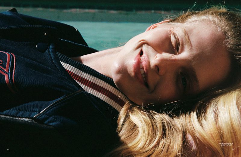 Close-up of a blonde woman lying down, smiling with eyes closed, wearing a dark blue varsity jacket with white and red striped collar.