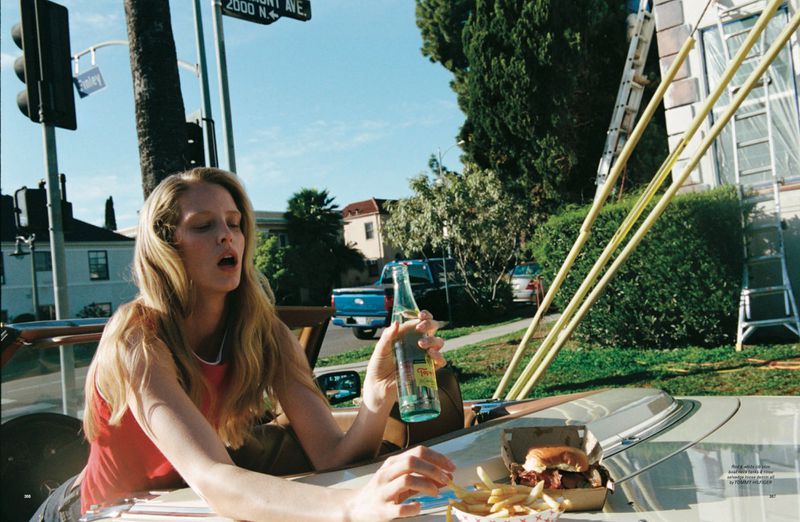 Woman eating fries and a burger in a car on a sunny day, with a traffic light and palm trees in the background, soda bottle in hand.