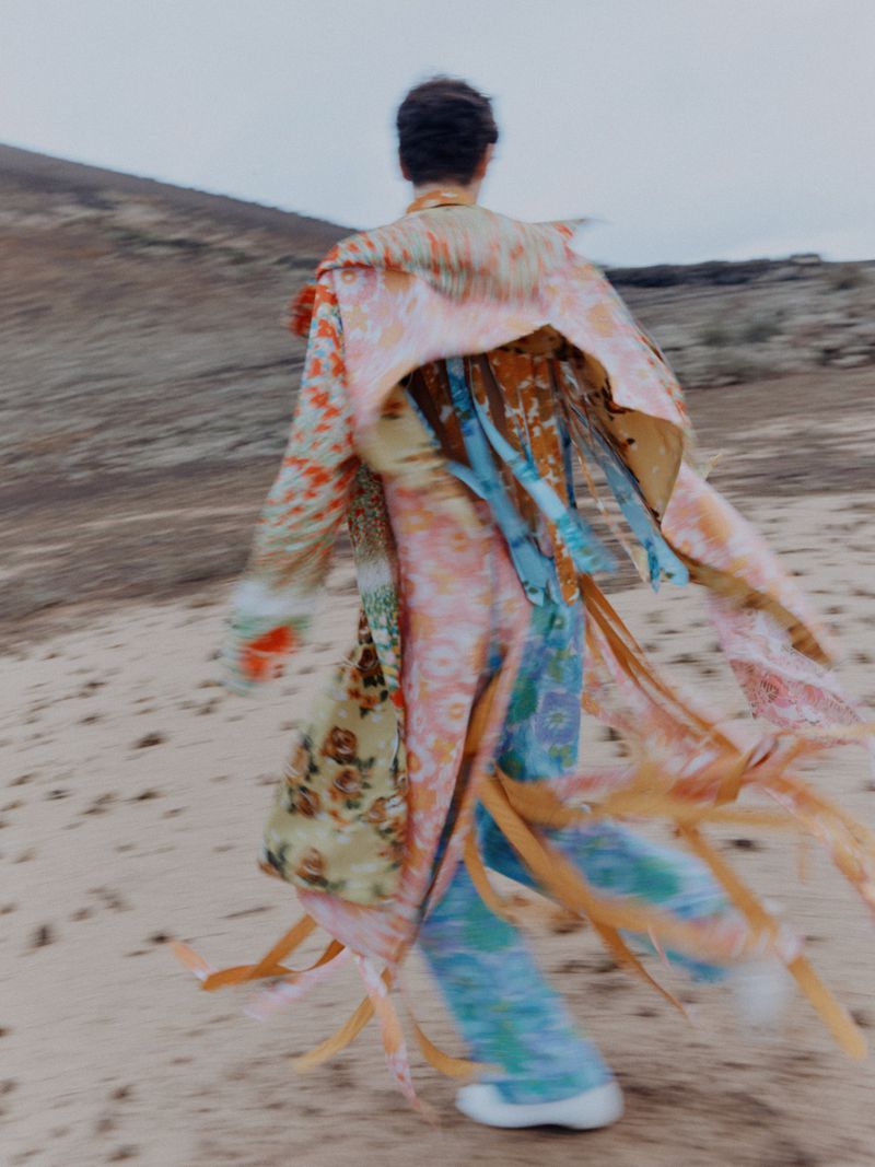 Person walks away in desert wearing a colorful patterned robe-like jacket with flowing ribbons and pants in a blurred, artistic style.