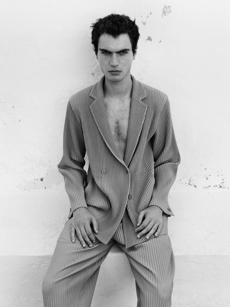 Monochrome portrait of a man in a pleated suit sitting against a weathered wall, captured in a fashion editorial style.