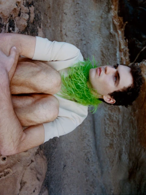 Young man poses crouched on a rock in front of a cave, wearing a light top and a bright green feathered boa around his neck.