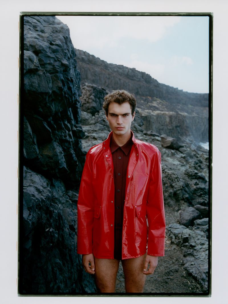 Male model in a red vinyl jacket and dark red shirt poses against a rocky cliff landscape with the ocean and sky in the background.