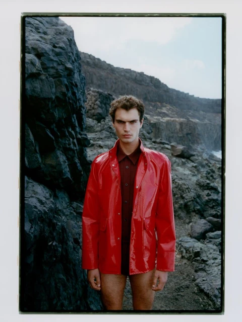 Male model in a red vinyl jacket and dark red shirt poses against a rocky cliff landscape with the ocean and sky in the background.