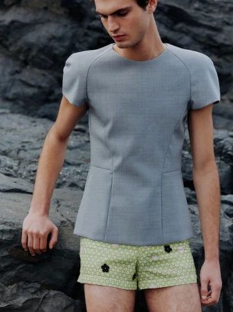 A young adult male model poses outdoors wearing a grey wool top and floral print shorts, against a dark rock formation backdrop.
