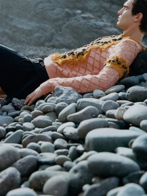 A fashionable adult male is resting on grey stones at a rocky beach wearing an ornate pink and gold sweater with black pants.