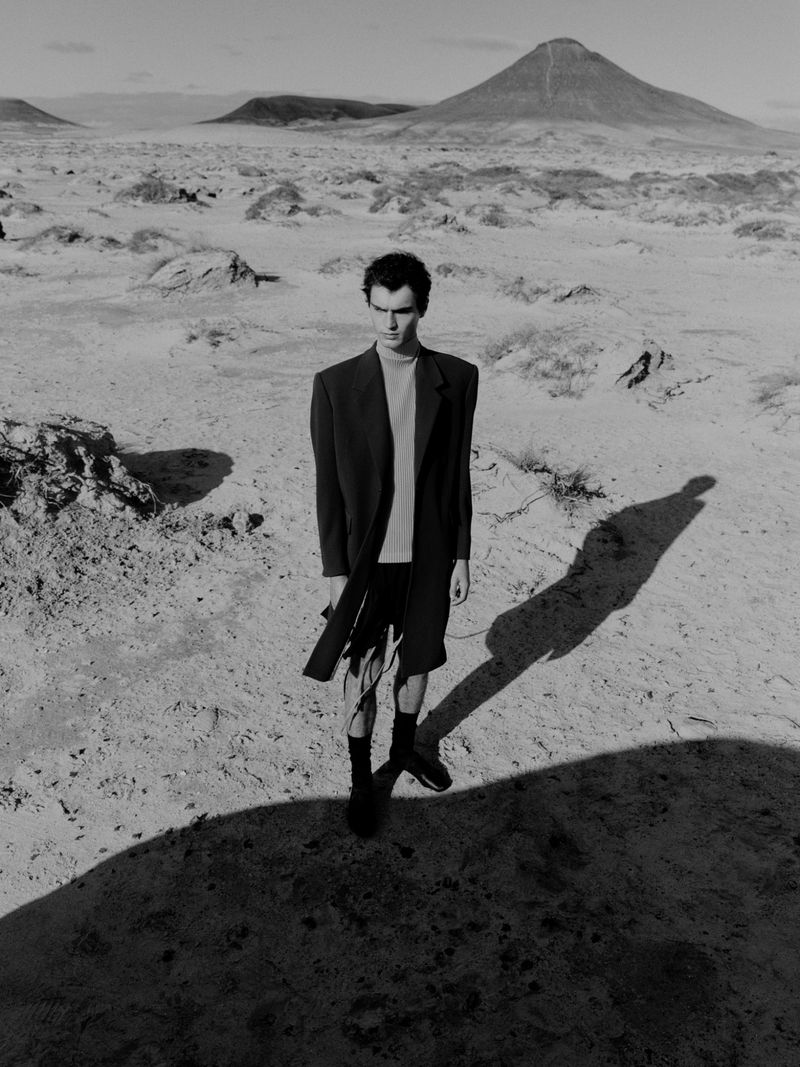 Black and white outdoor portrait of an adult male standing in a barren landscape with a volcano peak in the background, casting long shadow.