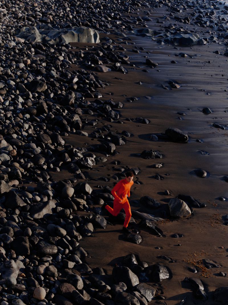 Person in orange running on rocky beach. A vibrant figure against a rugged coastal landscape with dark sand and large rocks near the water.