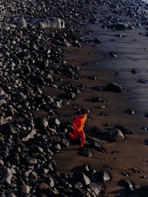 Person in orange running on rocky beach. A vibrant figure against a rugged coastal landscape with dark sand and large rocks near the water.