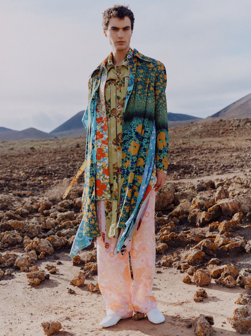 Fashion shot of a young adult man in a multicolor floral patterned shirt and pants standing in a rocky landscape, on a sunny day.