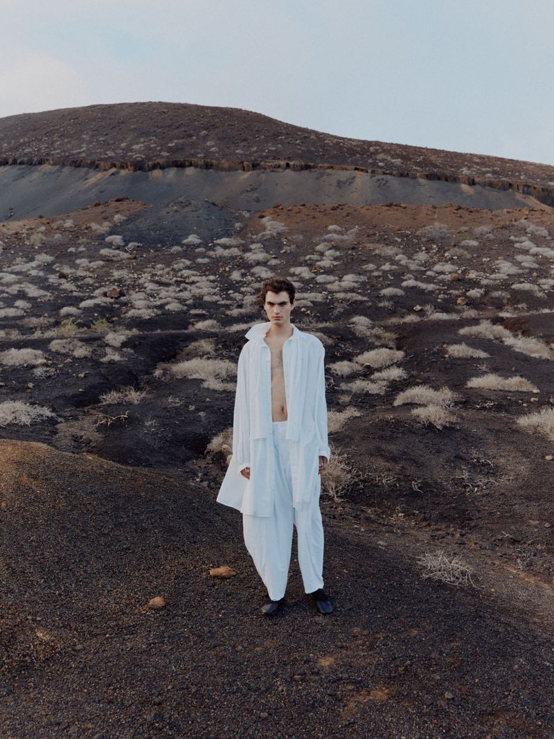 An adult male stands in an open white shirt and pants on a barren, hilly landscape under a clear sky in a fashion-style portrait.
