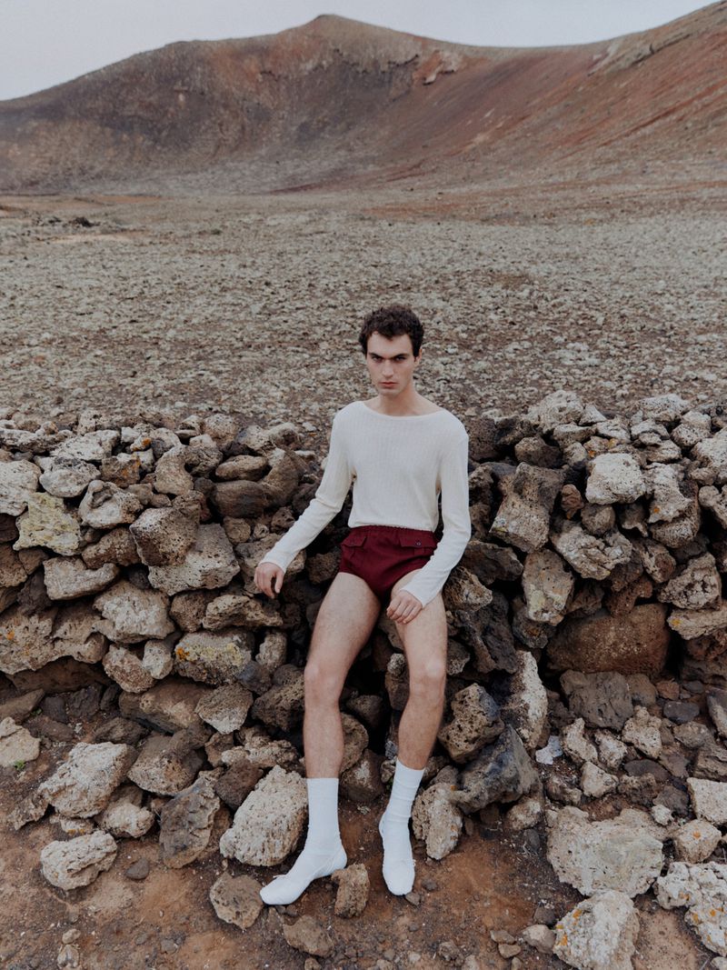 Young man posing outdoors in a cream long-sleeve shirt, maroon shorts, and white socks, sitting on a stone wall near a mountain.