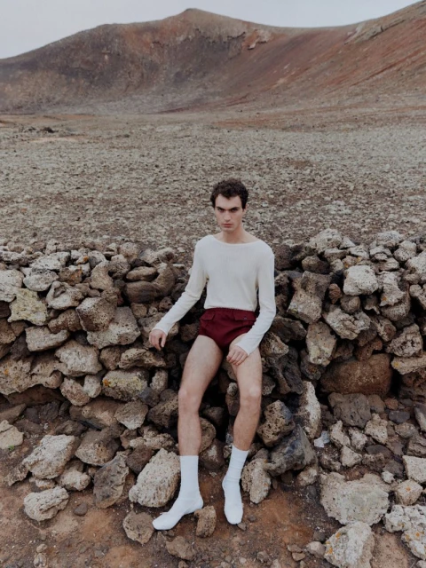 Young man posing outdoors in a cream long-sleeve shirt, maroon shorts, and white socks, sitting on a stone wall near a mountain.