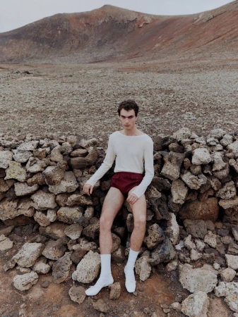 Young man posing outdoors in a cream long-sleeve shirt, maroon shorts, and white socks, sitting on a stone wall near a mountain.