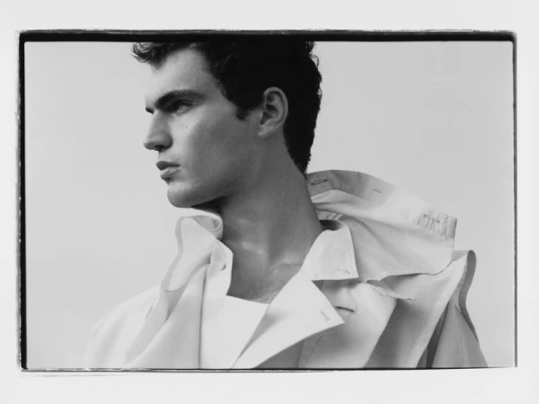 Monochrome portrait of a man with dark hair wearing a white collared shirt and jacket, looking left against a white backdrop.