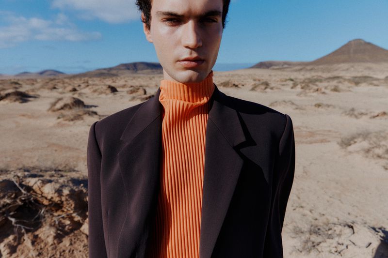 A fair-skinned man in a black blazer and orange turtleneck sweater poses in a desert landscape with mountains and blue sky in the background.