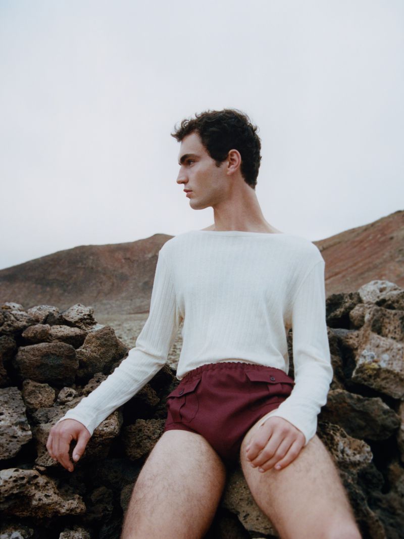 A young adult male model poses in front of a rocky landscape, wearing a white long-sleeve shirt and maroon shorts in a fashion shot.