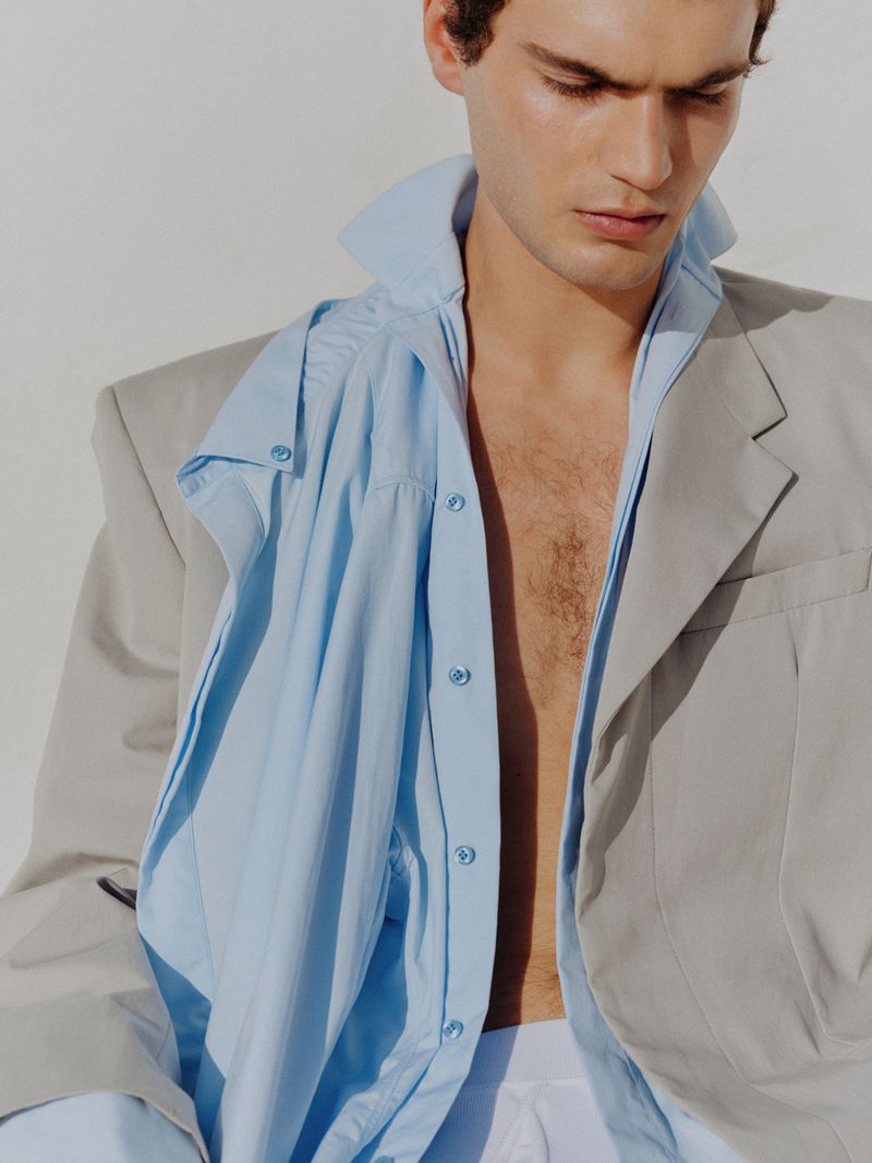 Close-up portrait of a male model wearing an unbuttoned blue shirt and a grey jacket against a white background