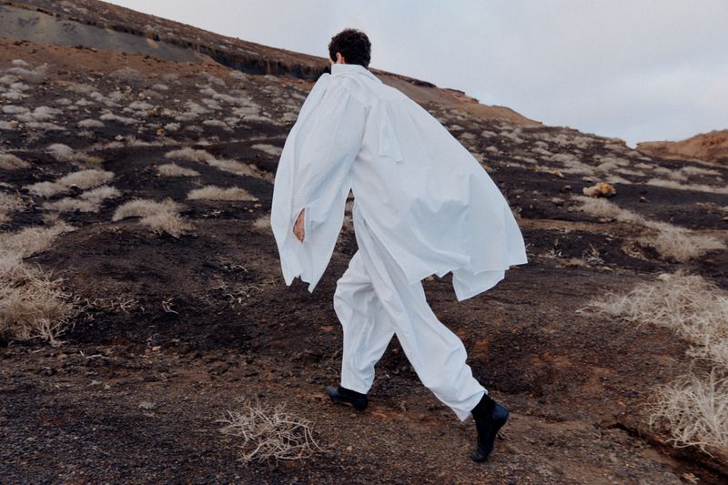 A solitary adult walks in a billowing white outfit across a stark, rocky landscape under an overcast sky, creating a serene, surreal scene.