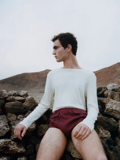 Model Lars Jammaers wearing a white long-sleeve top and burgundy shorts posing outdoors on rocky terrain for The Greatest Magazine.
