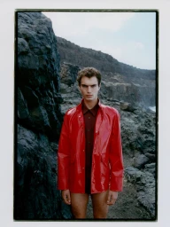 Model wearing a glossy red jacket standing against a rocky coast and dark stone cliff under an overcast sky.