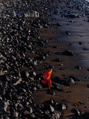 Model in bright orange clothing running across a dark rocky beach shoreline shot from an elevated aerial perspective