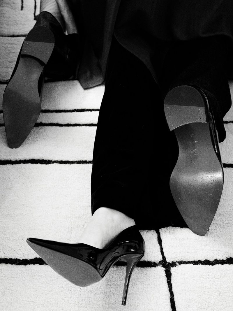 Close-up of woman in black pants and shiny black high heels, with a cream and black geometric rug visible underneath.