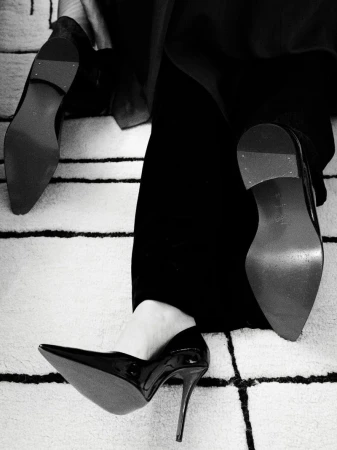 Close-up of woman in black pants and shiny black high heels, with a cream and black geometric rug visible underneath.