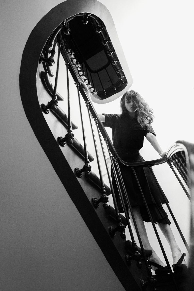 Monochrome portrait of a woman in a dark dress on a decorative staircase, captured from a low angle, emphasizing architectural details.